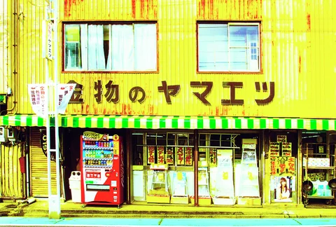 A yellow shop with green-striped awnings displays Japanese signage above glass doors and a red vending machine.