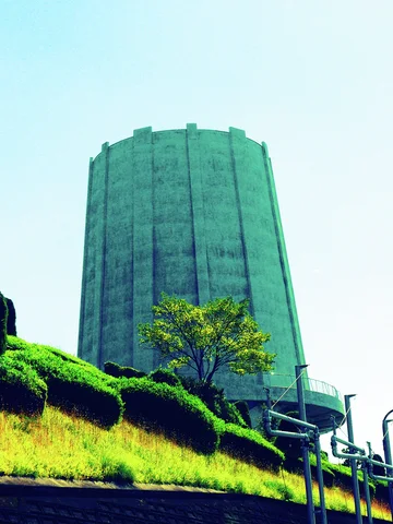 A tall green cylindrical structure rises above a grassy bank, trimmed hedges, and a small tree.