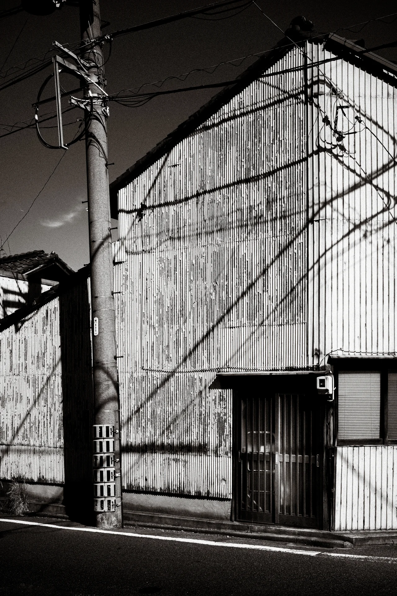 Utility wires cast angular shadows across a corrugated building beside a concrete pole in black and white.