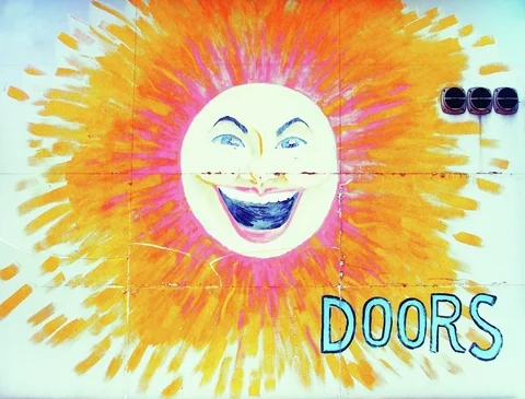 A smiling face surrounded by orange and pink rays decorates a pale wall beside blue Doors lettering.
