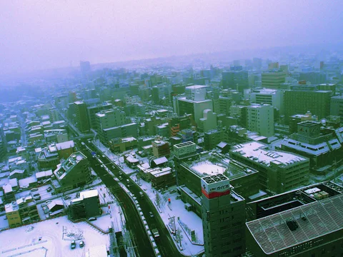A dark curving road divides snow-covered city blocks beneath a lavender hazy sky.