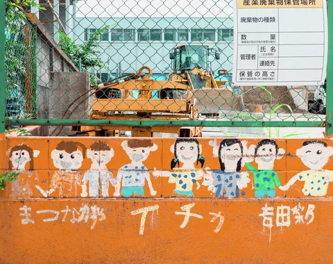 Painted children line an orange barrier below chain-link fencing enclosing yellow machinery and a posted notice.