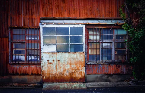 A rust-stained metal door stands between barred windows on a reddish corrugated facade with climbing greenery.