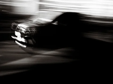 A dark car passes through strong horizontal motion blur along a street in black and white.