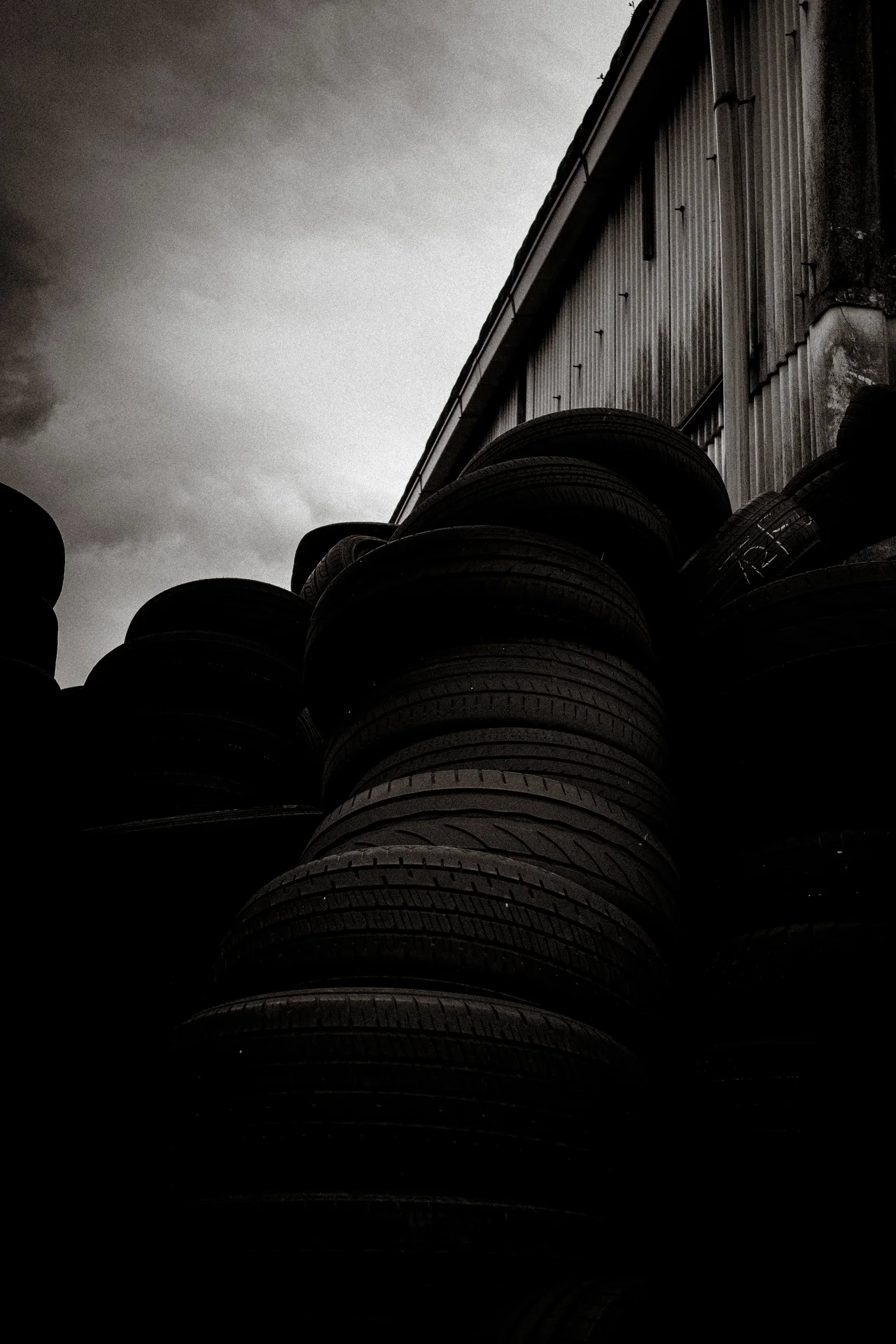 Tall stacks of tires rise beside a corrugated building under a cloudy sky in black and white.
