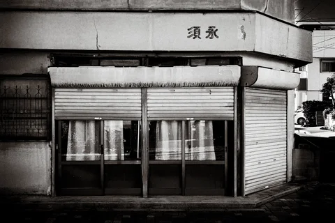 Curtained glass doors stand beneath partly lowered shutters on a weathered corner shop in monochrome.