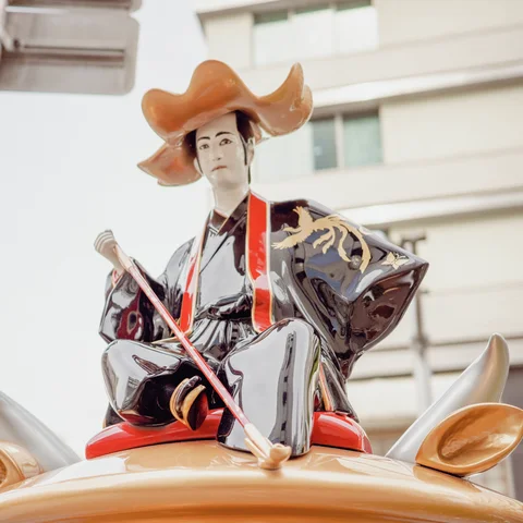A seated robed figure sculpture wears a broad orange hat and holds a slender staff.