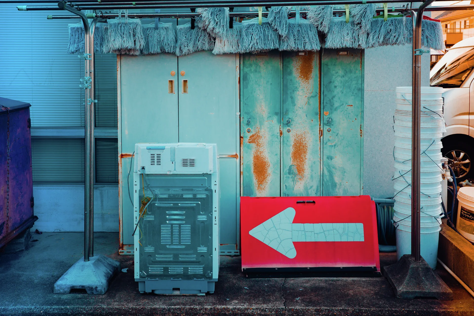 A red left-arrow sign rests beside stacked crates against weathered turquoise doors streaked with rust.