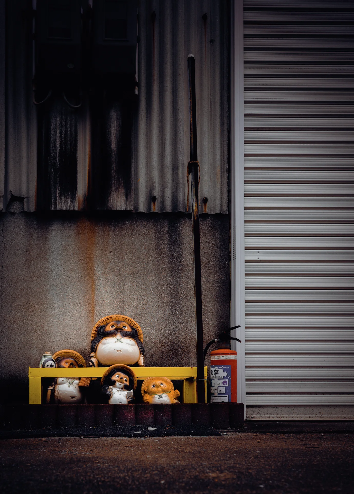Small rounded animal figurines cluster beneath a yellow bar beside a closed shutter and stained wall.