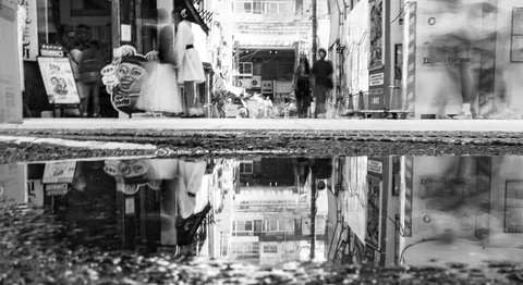 A puddle reflects storefronts and passing pedestrians beneath a rough pavement edge in black and white.