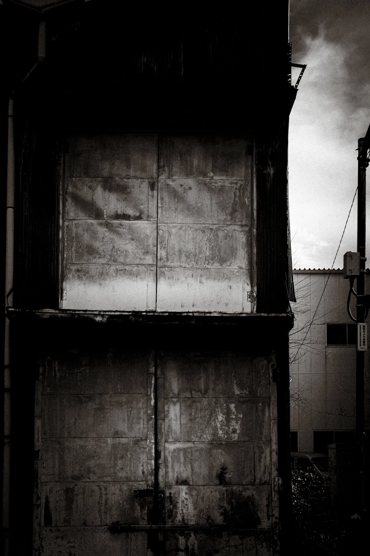 Two pairs of weathered closed shutters cover a dark facade beside a narrow strip of sky.