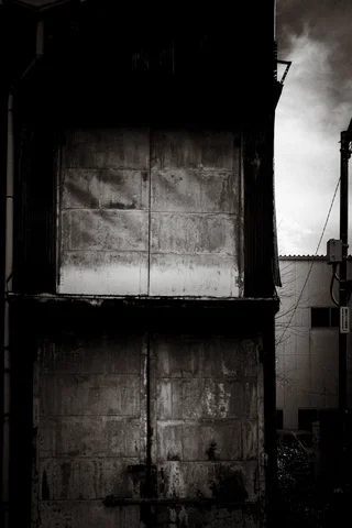 Two pairs of weathered closed shutters cover a dark facade beside a narrow strip of sky.