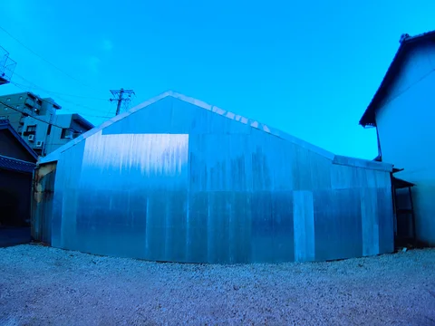 A curved blue corrugated enclosure occupies a gravel plot between neighboring buildings beneath clear sky.
