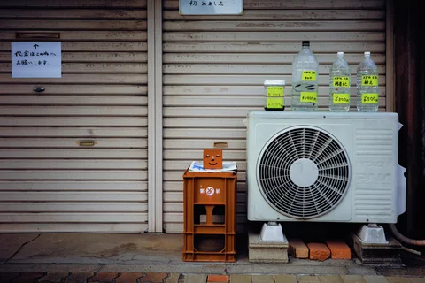Labeled bottles rest atop an outdoor air conditioner beside an orange crate and closed shutters.