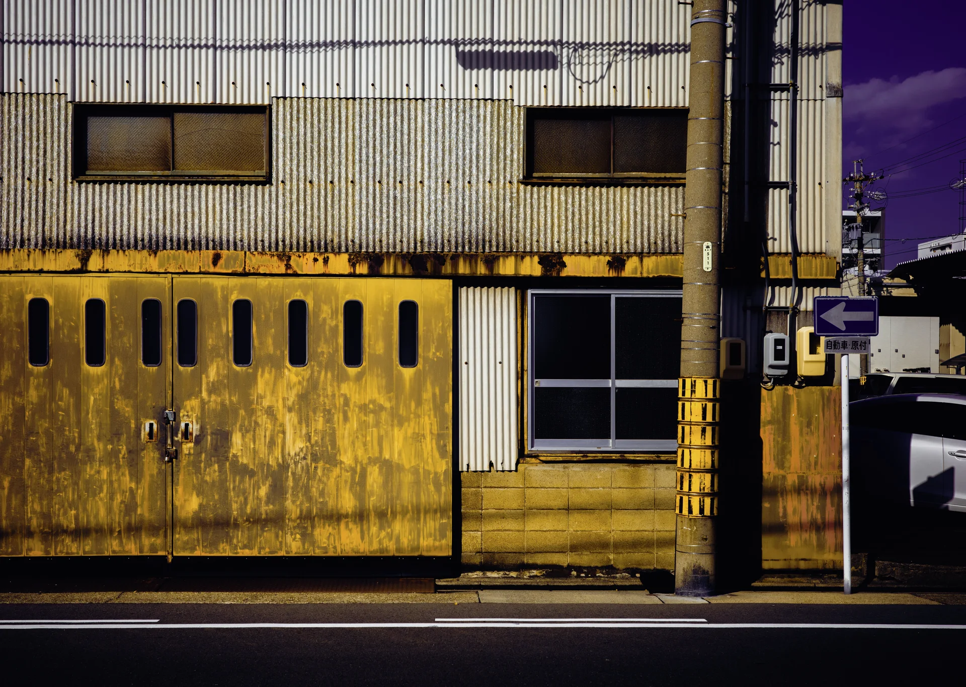 Rust-stained yellow doors line a corrugated industrial building beside a pole and blue left-arrow sign.