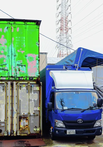 A small blue truck stands beside stacked weathered shipping containers beneath a distant electricity pylon.