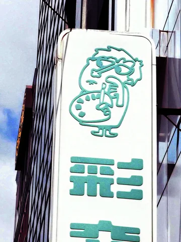 A cartoon artist with glasses and a palette decorates a tall white sign above turquoise Japanese characters.