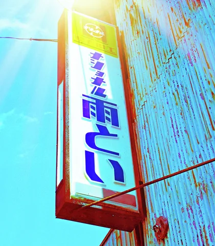 Sunlight flares above a narrow projecting sign with blue Japanese lettering beside rust-streaked turquoise siding.
