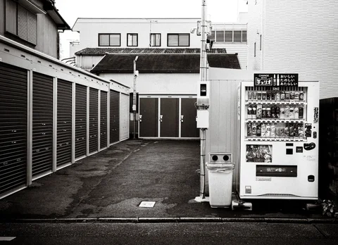 A vending machine stands beside a paved courtyard lined with closed shutters in black and white.