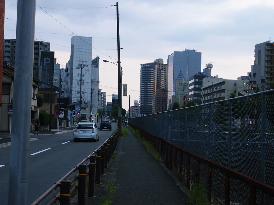 Cars travel along a city road beside a fenced narrow sidewalk leading toward tall office buildings.