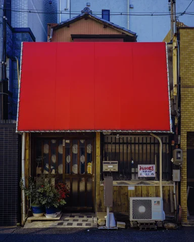 A large blank red sign dominates a narrow storefront with weathered panels and potted plants below.