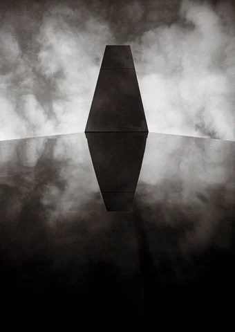 A dark tapered geometric structure meets its reflection amid cloudy light in black and white.