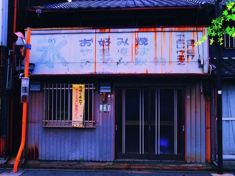A rust-streaked pale shop sign hangs above a barred window and dark sliding entrance doors.