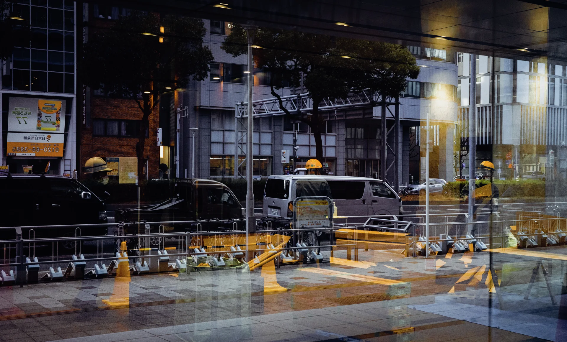 Glass reflections overlap parked vehicles, street railings, and yellow posts outside a large building.