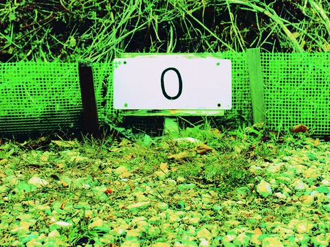 A white rectangular marker bearing a black zero stands against green mesh beside gravel and tall grass.