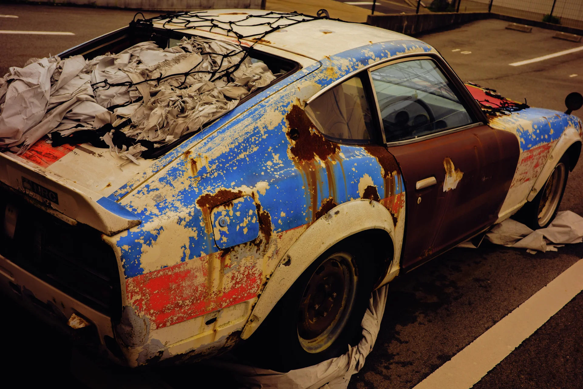 Peeling blue paint and extensive rust cover a sports car piled with fabric beneath its damaged rear window.