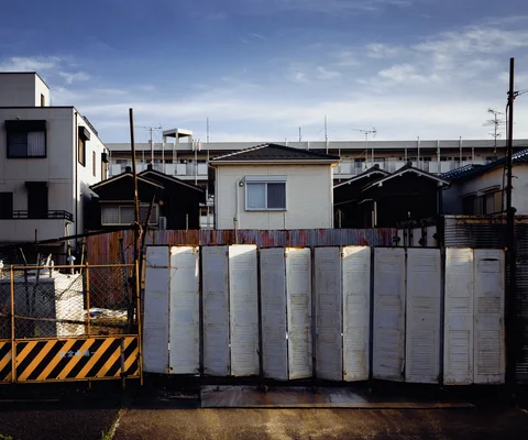 Pale construction panels and a striped barrier enclose a site between small houses beneath blue sky.