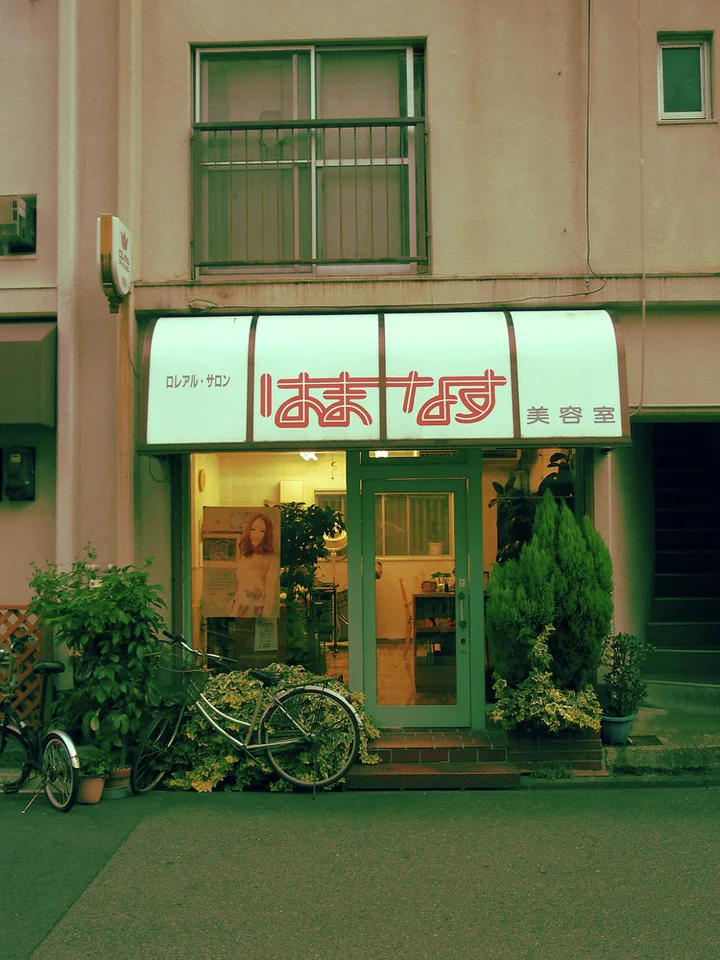 A small shop has golden glass doors beneath a pale awning with bicycles and shrubs outside.