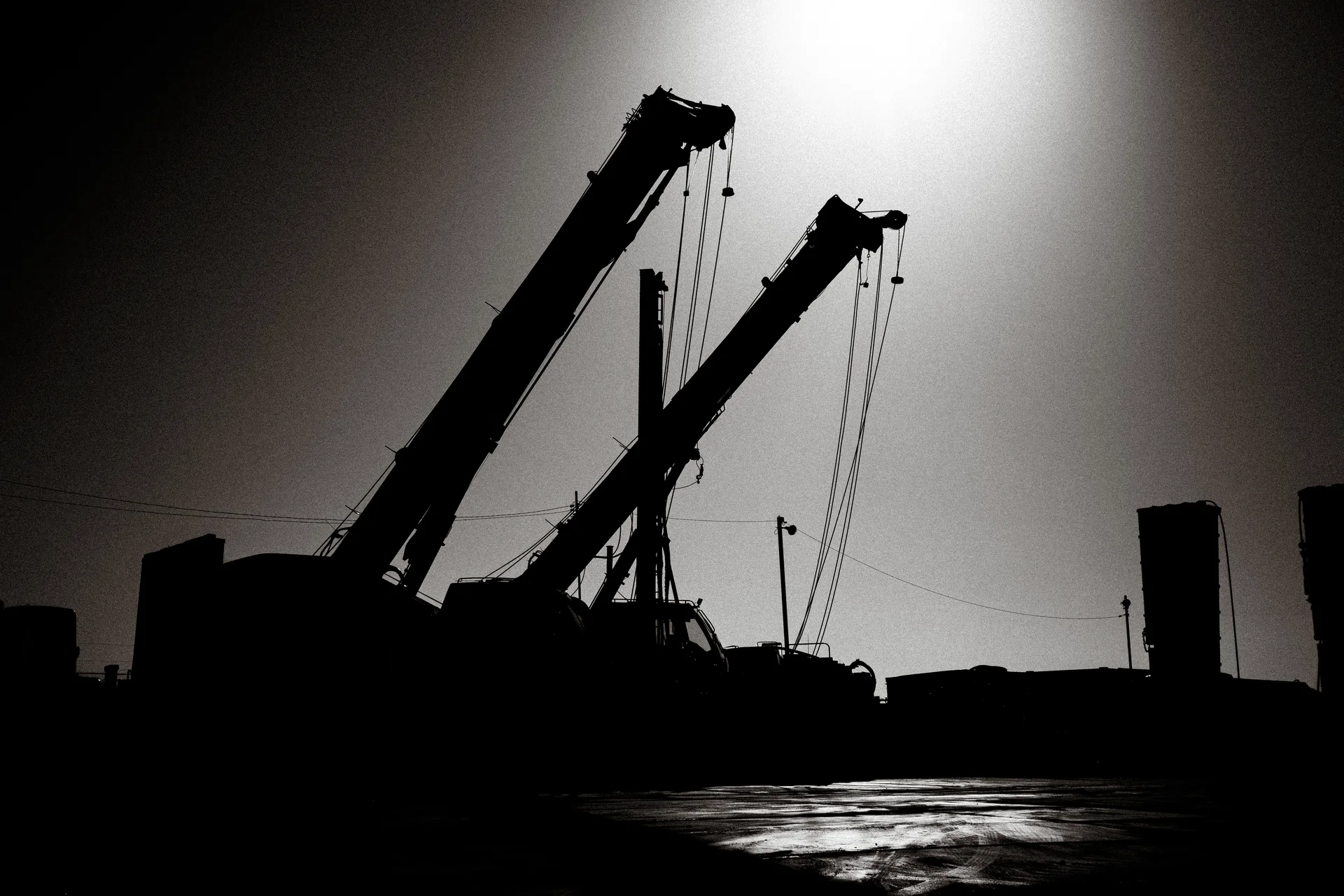 Two crane booms and hanging cables form dark silhouettes above reflective ground in black and white.