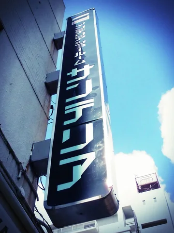 A tall dark projecting sign carries pale Japanese lettering beside a building against bright blue sky.