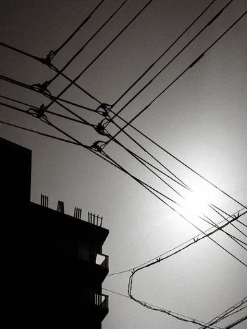 Overhead utility wires crisscross the bright sun beside a silhouetted building in black and white.