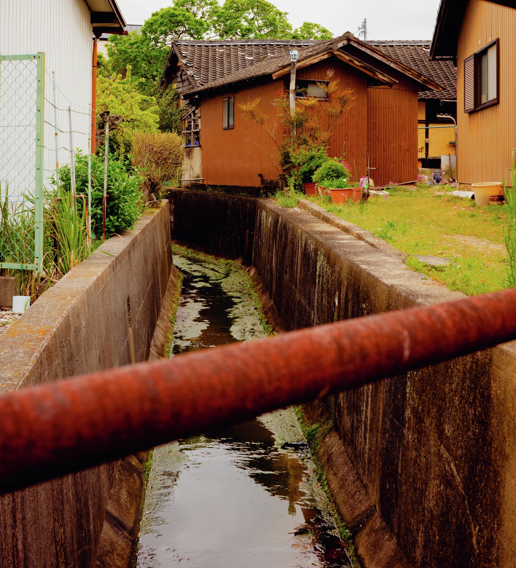 A rusty pipe crosses a narrow concrete drainage channel beside grass and orange-sided houses.
