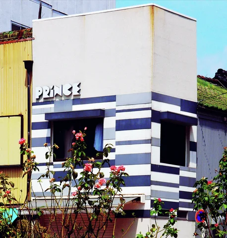 Pink flowers rise before a small white building striped in dark blue around square recessed windows.