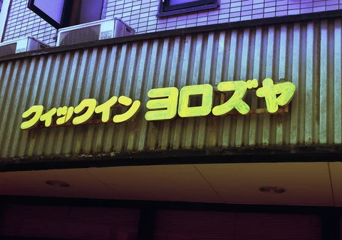 Raised yellow Japanese lettering spans a weathered corrugated facade beneath pale square tiles.