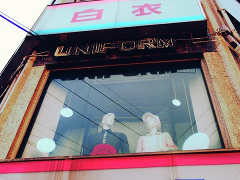 Two pale mannequins stand behind a large upstairs shop window beneath weathered uniform lettering.