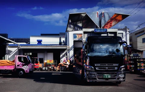 A large dark truck and a smaller pink truck stand before industrial buildings beneath angular roof structures.