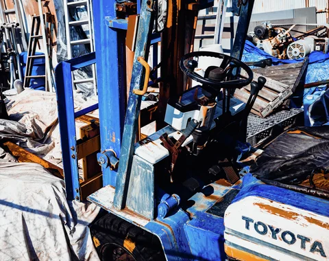 A worn blue forklift sits among ladders, tarps, timber, and machinery in a crowded outdoor work area.