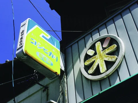 A yellow projecting sign hangs beside a large circular character emblem on a corrugated wall.