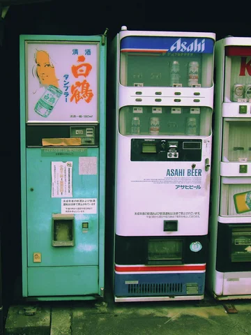 Green and white vending machines stand together beneath a dark overhang beside a narrow illustrated cabinet.