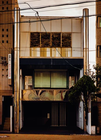 A narrow pale building has dark horizontal awnings, upper window grilles, and a recessed ground-floor entrance.