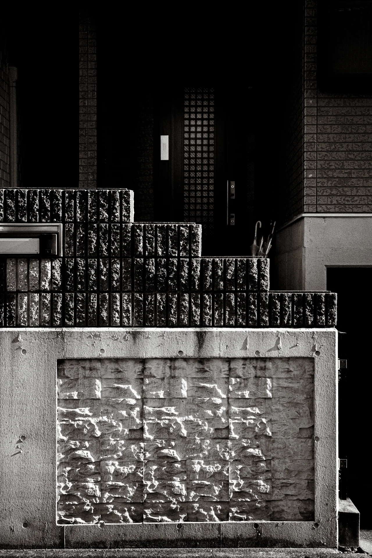 A patterned concrete wall fronts a shadowed entrance behind stepped dark blocks in black and white.