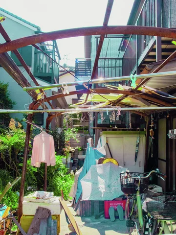 Laundry hangs beneath a rusty curved framework over a cluttered courtyard with bicycles and plants.