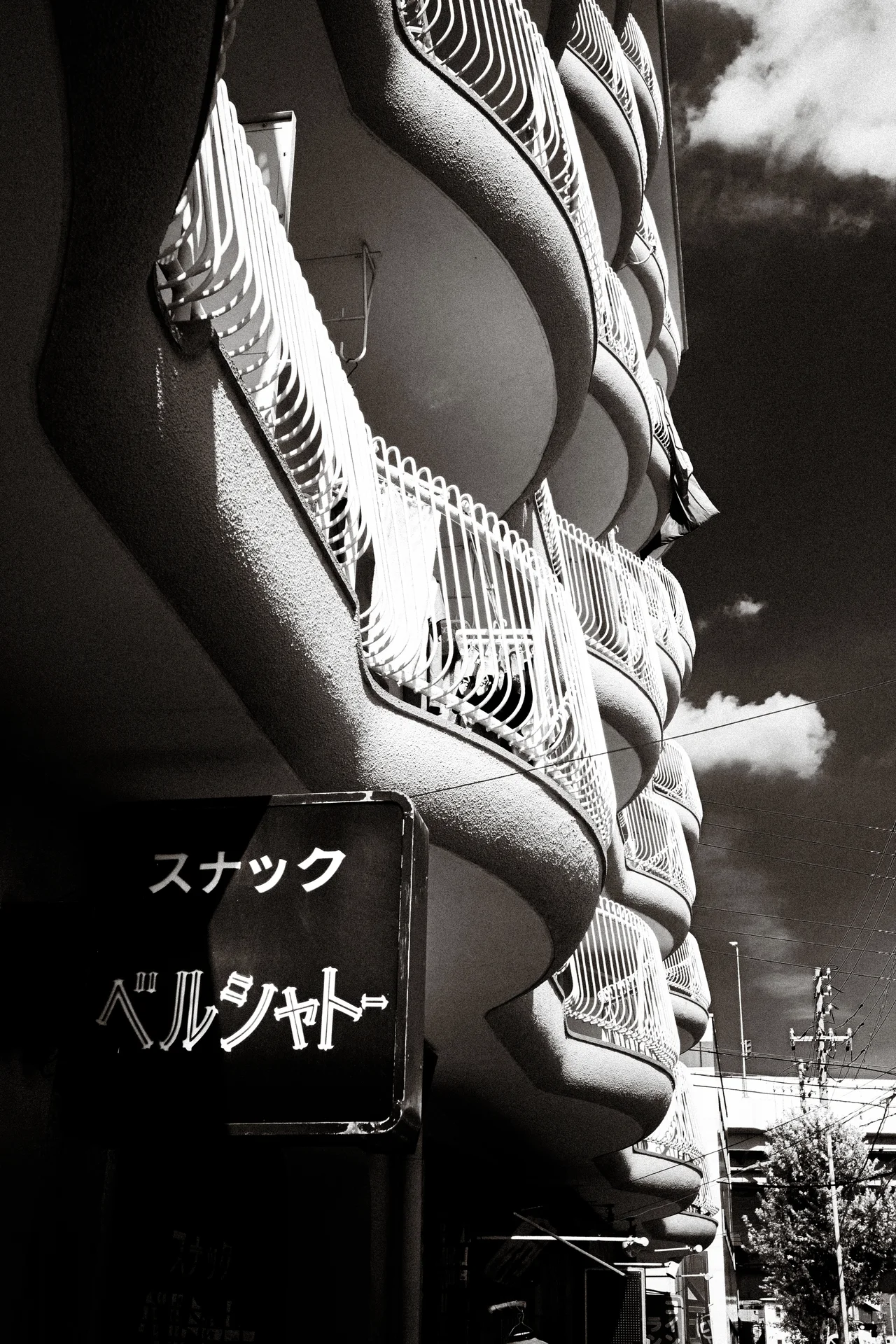 Rounded concrete balconies with decorative curved railings rise above a projecting sign in black and white.
