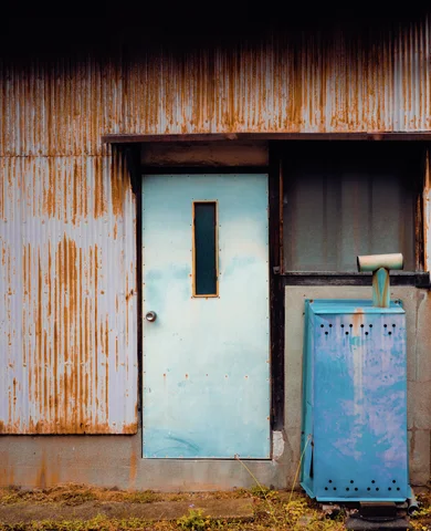 A pale blue door and turquoise metal box stand against rust-streaked corrugated siding.