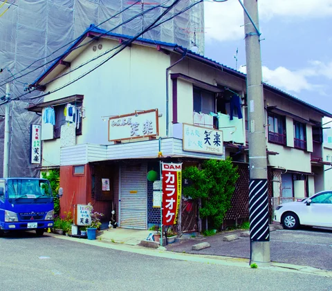 A pale corner building has hanging laundry, faded signs, and a red karaoke banner beside a utility pole.