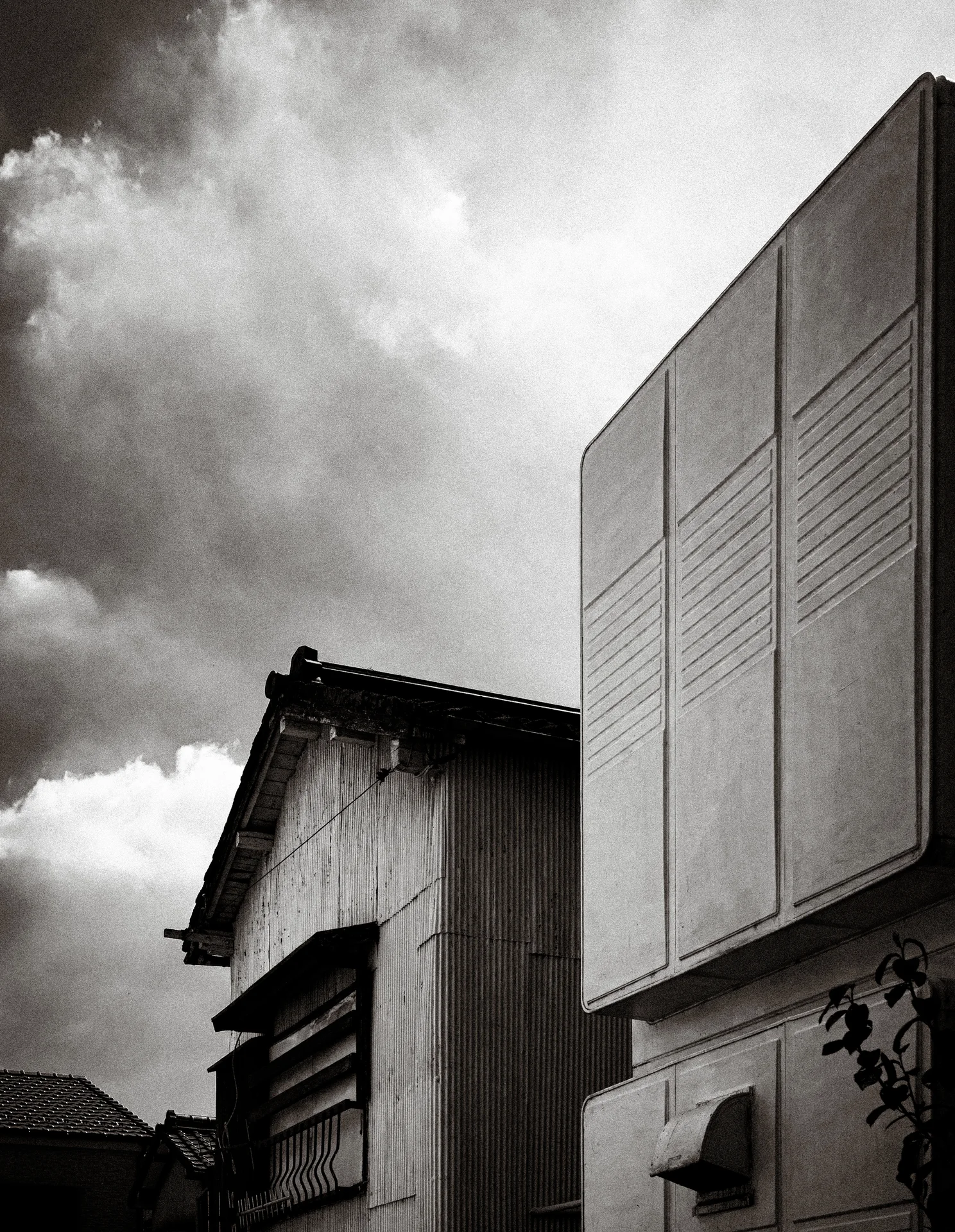 A tall paneled wall rises beside a low corrugated building beneath textured clouds in black and white.